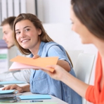 junge Frau im Büro reicht einer weiteren jungen Frau eine Mappe weiter, im Hintergrund ein junger Mann